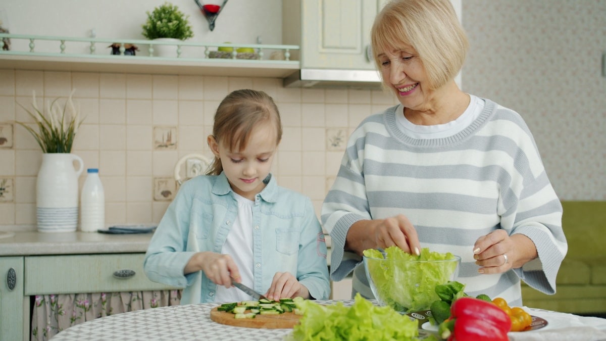 Granddaughter and grandmother are cooking salad cutting vegetables then doing high-five and laughing enjoying time together. People and house concept.